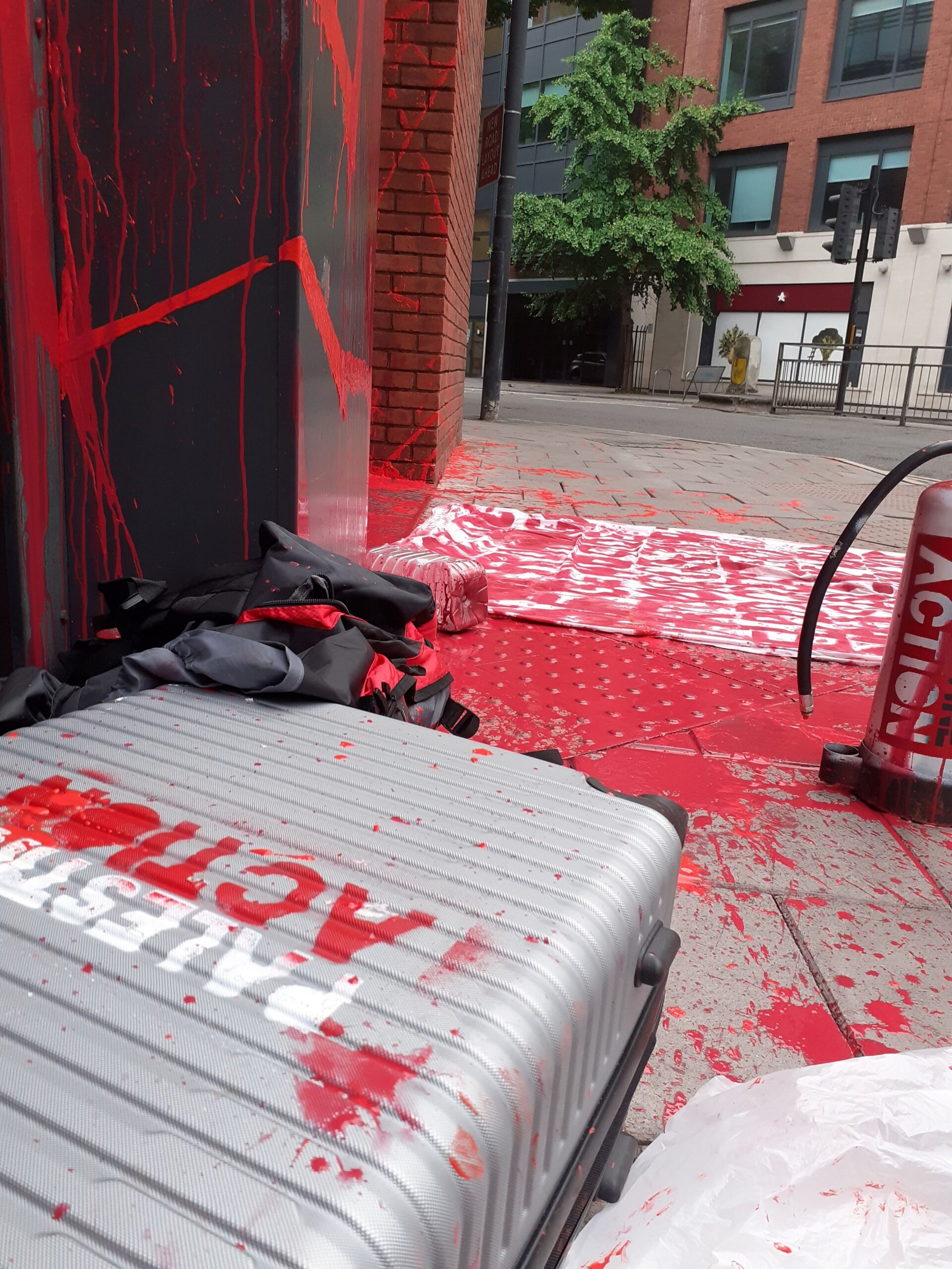 Silver suitcase with Palestine Action spraypainted on it in white and red. Red paint splatters cover the pavement and walls, next to a fire extinguisher. 