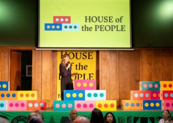 A woman stands on stage with 'House of the People' on a board above and on a banner behind her. A crowd of people sit watching.