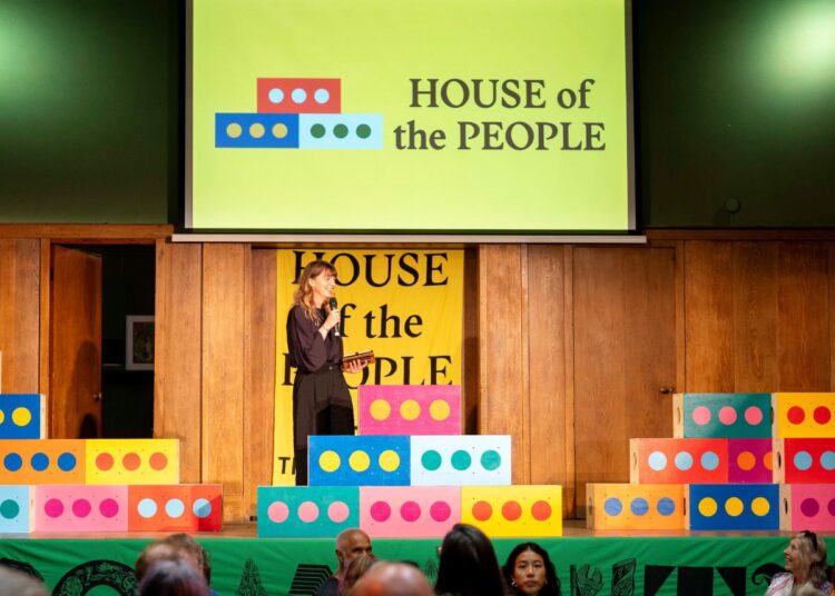 A woman stands on stage with 'House of the People' on a board above and on a banner behind her. A crowd of people sit watching.
