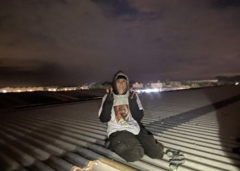 Palestinian Martyrs for Justice activist sits on top of Moog's roof making peace signs and wearing a T-shirt with the name and face of a Palestinian martyr.