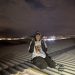 Palestinian Martyrs for Justice activist sits on top of Moog's roof making peace signs and wearing a T-shirt with the name and face of a Palestinian martyr.