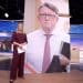 Laura kuenssberg standing in front of a giant picture of Peter Mandelson