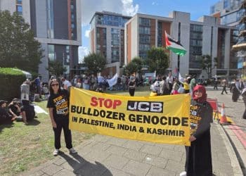 Two protesters hold a yellow banner outside DSEI reading: "Stop JCB bulldozer genocide in Palestine, India & Kashmir".