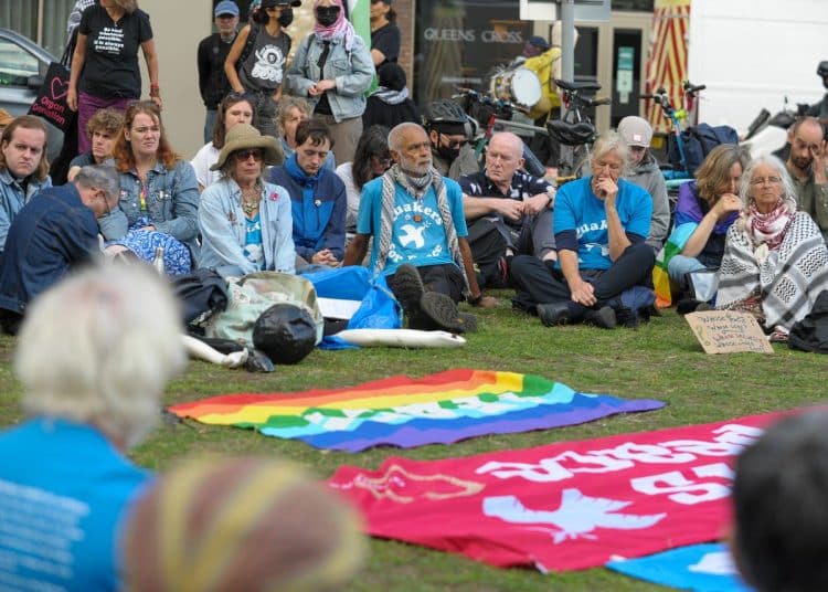 A group of Quakers sit together in pale blue t-shirts on the grass outside DSEI in silent protest.