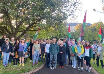 A large group of walkers stand together under a large tree waving Palestine flags Gaza