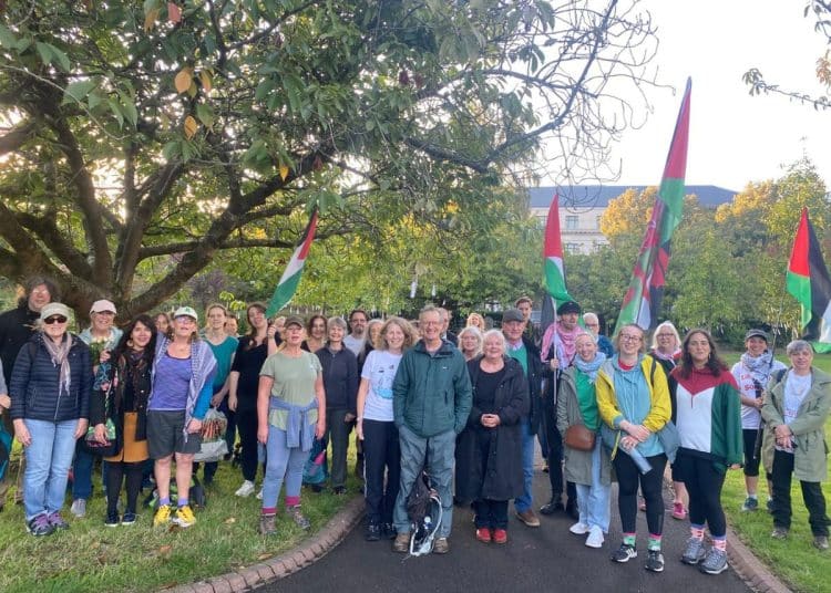 A large group of walkers stand together under a large tree waving Palestine flags Gaza