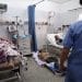 Women and man in hospital beds, with one patient on the floor and a medic in scrubs walking towards them.