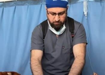 Turkish doctor Taner Kamaci in scrubs treating a child on a hospital bed hooked up to an IV drop, with blue hospital curtains behind them.