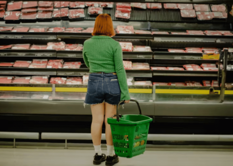 Woman stands infront of a supermarket shelf of meat holding a basket