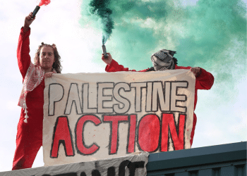 Palestine Action activists stand holding a 'Palestine Action' banner and holding coloured smoke flares in their free hands