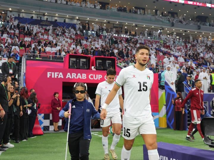 A child player escort accompanies a player at the Arab Cup. The child is wearing dark glasses and carrying a white cane.