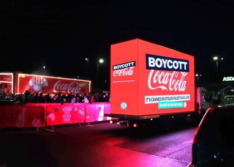In the foreground, a bright red truck with Boycott Coca-Cola written on it. In the background, the Coca-Cola Christmas truck