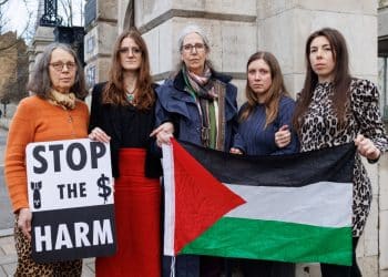 Five women stand in front of a stone building holding a Palestine flag and a placard saying Stop The Harm with images of a bomb and a dollar sign