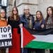 Five women stand in front of a stone building holding a Palestine flag and a placard saying Stop The Harm with images of a bomb and a dollar sign