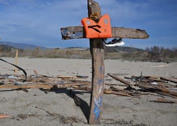A cross made with driftwood from the Cutro shipwreck. A lifejacket and a training shoe hang from the cross