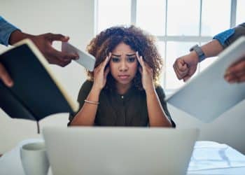 Image illustrating workplace stress. A tired looking woman sits behind a laptop as hands holding a phone, a watch, a report, a book, intrude from the sides