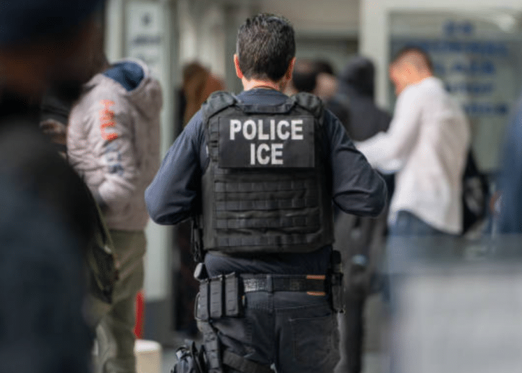 ICE officer has his back to the camera, capturing only his bulletproof vest with 'Police ICE' on it