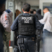 ICE officer has his back to the camera, capturing only his bulletproof vest with 'Police ICE' on it