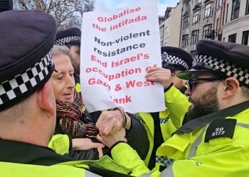 Peter Tatchell surrounded by cops and holding a placard saying Globalise The Intifada. Non-violent Resistance. End Israel's Occupation Of Gaza & West Bank