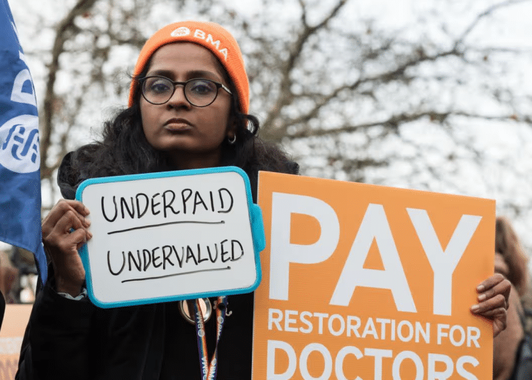 Doctors strike outside St Thomas' Hospital in London on 17 December 2025. A woman holding two placards, one says 'Underpaid, undervalued' and the other 'Pay restoration for doctors'.