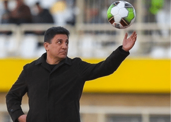 Iran national football team coach, Amir Ghalenoei, throws a football in the air with one hand while the other rests confidently in his pocket. He resembles a strong statue and stares at the ball in mid-air.