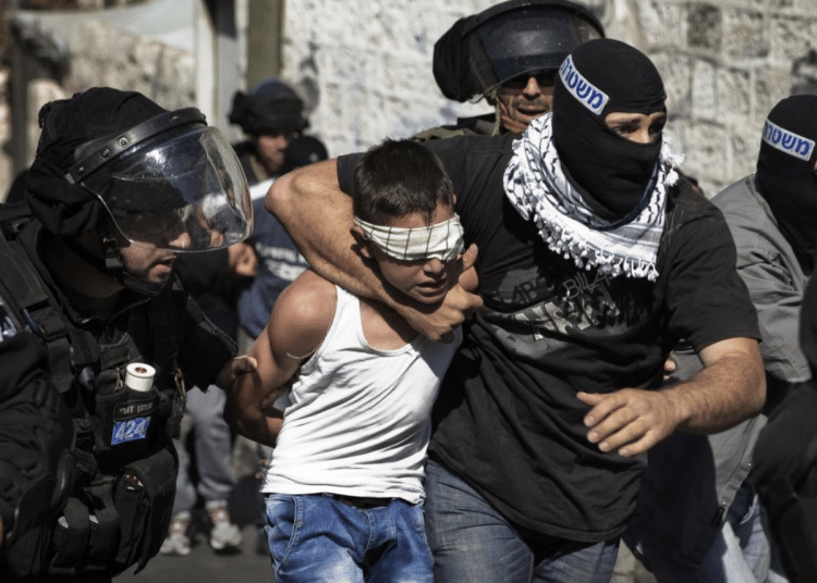 A child in Palestine, a boy, is gripped around his neck by an IOF member while blindfolded. His hands are held behind his back
