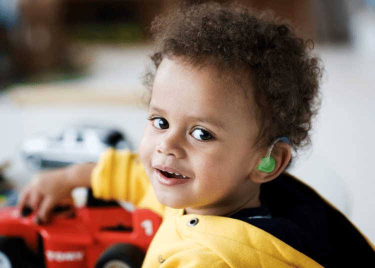 A deaf young boy with curly hair turns to smile at the camera with one hand on his plastic toy car. His ear shows his colourful hearing aid.