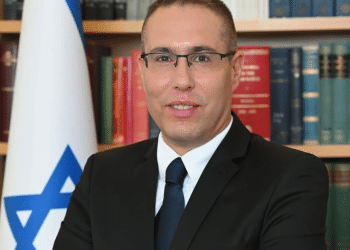 Eli Hazan, Benjamin Netanyahu's comms director smiles in a corporate headshot with the Israel flag and a packed bookshelf behind him