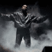 Kanye West on a stage dressed in black surrounded by smoke with his arms outstretched