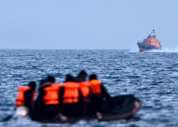 A group of people wearing life jackets sit on an inflatable boat in the middle of the sea. A larger vessel in the distance sails towards them