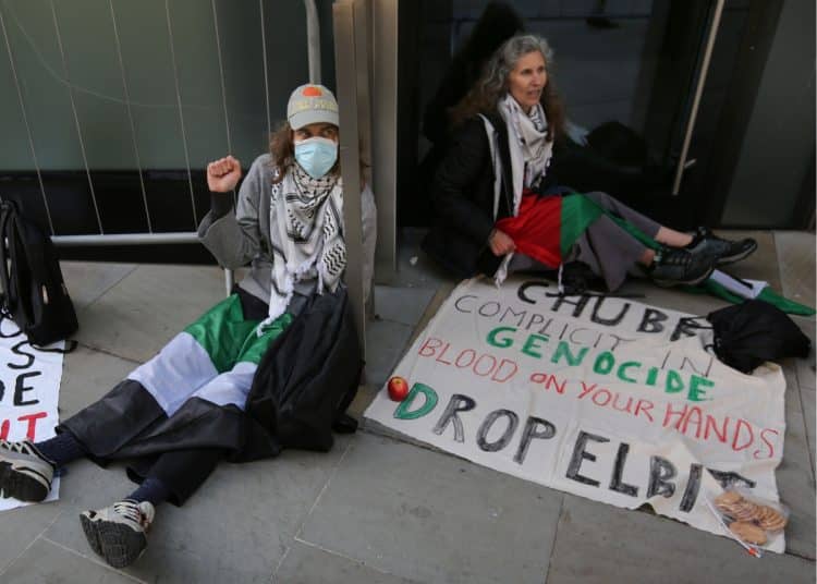 Two protesters sit, locked-on, outside the London HQ of Chubb Insurance. A banner reads: Chubb Complicit In Genocide Blood On Your Hands Drop Elbit