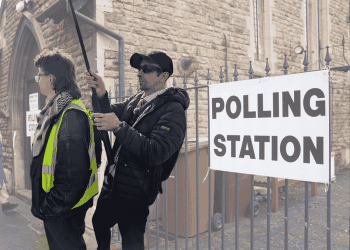 The background is an image of a polling station. In the foreground there's an image of Lee Scheresy standing weirdly close to a protest steward. He has a hat and sunglasses on and is holding a camera on a long pole - auditor