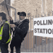 The background is an image of a polling station. In the foreground there's an image of Lee Scheresy standing weirdly close to a protest steward. He has a hat and sunglasses on and is holding a camera on a long pole - auditor