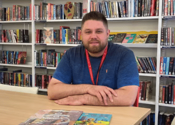 Jakob Williamson sat at a table at what appears to be a library in Wakefield, with his arms folded and rested on the table with book shelves behind him