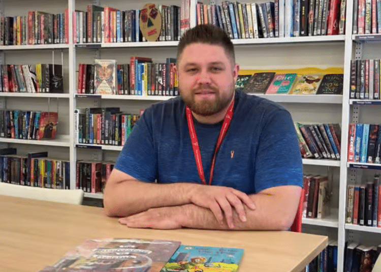 Jakob Williamson sat at a table at what appears to be a library in Wakefield, with his arms folded and rested on the table with book shelves behind him