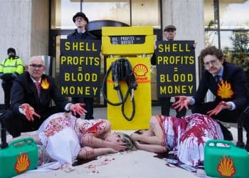 Two people lie on the ground covered in fake blood. Suited men with fake blood on their hands are behind them. On either side of a fake petrol pump, signs read Shell Profits = Blood Money