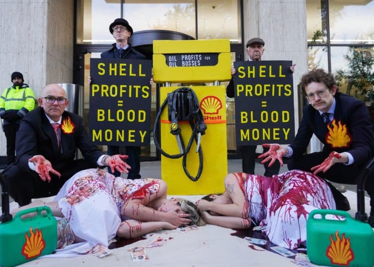 Two people lie on the ground covered in fake blood. Suited men with fake blood on their hands are behind them. On either side of a fake petrol pump, signs read Shell Profits = Blood Money