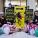 Two people lie on the ground covered in fake blood. Suited men with fake blood on their hands are behind them. On either side of a fake petrol pump, signs read Shell Profits = Blood Money