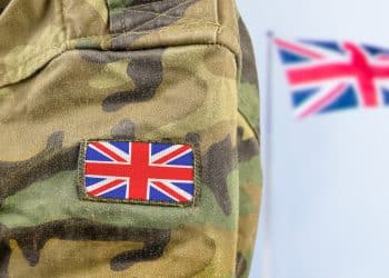 Close up of a soldier's arm in camo with a union flag badge. A union flag is flying in the background. Defence Universities Alliance