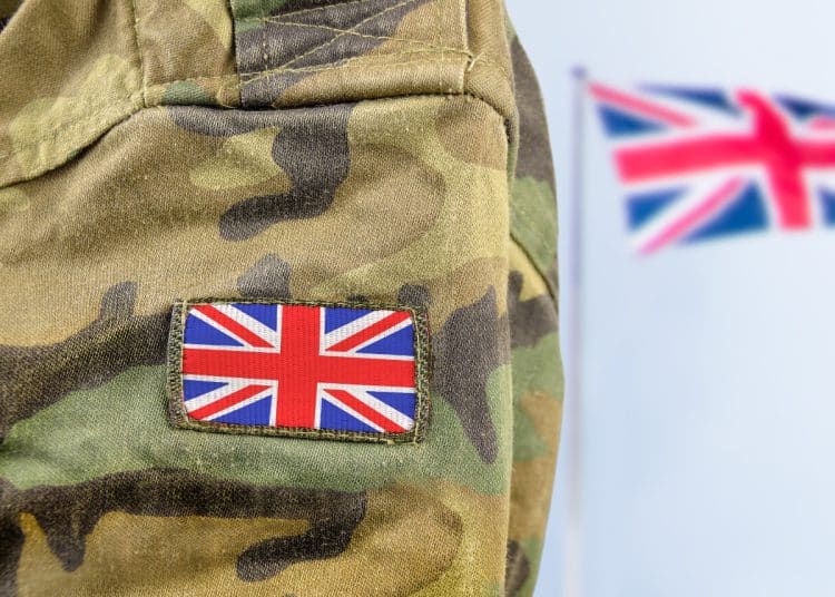 Close up of a soldier's arm in camo with a union flag badge. A union flag is flying in the background. Defence Universities Alliance