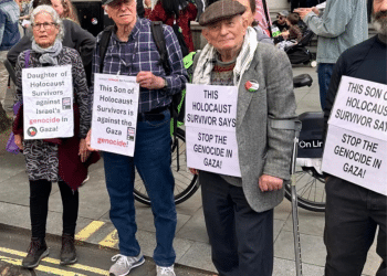 Holocaust survivor Stephen Kapos (right) and descendants of Holocaust survivors take part in an April 13, 2024 London protest against the Gaza genocide. He wears a flat cap and wool jumper underneath a wool blazer with one arm in a walking stick with arm support. On his torso is a poster that reads: ‘This Holocaust survivor says stop the genocide in Gaza’.