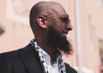 Majid Freeman, wearing sunglasses, smiles and looks into the distance. The stylish photograph captures his side profile.