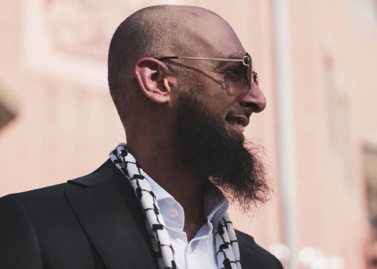 Majid Freeman, wearing sunglasses, smiles and looks into the distance. The stylish photograph captures his side profile.