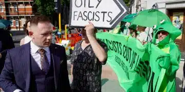 A suited man walks past protesters outside the ARC conference in London. A sign says Oily Fascists