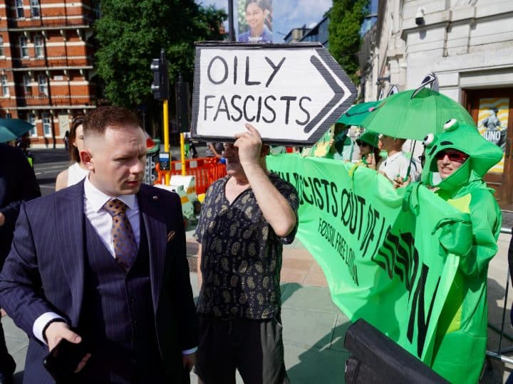 A suited man walks past protesters outside the ARC conference in London. A sign says Oily Fascists