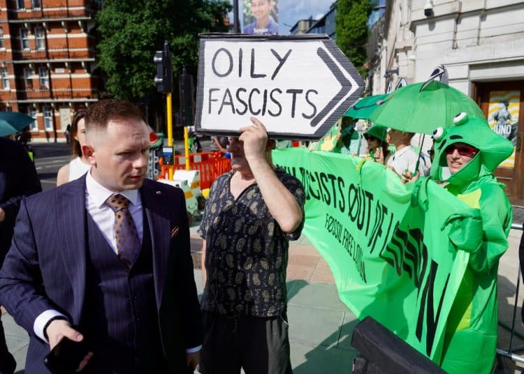 A suited man walks past protesters outside the ARC conference in London. A sign says Oily Fascists