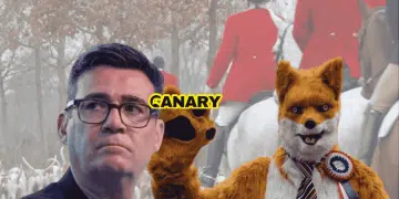 An image of the hunt and hounds in the background, wearing the traditional red coats. In the foreground on the left is Robert Pownall dressed as a fox. He is wearing a red, blue and white rosette and is waving at the camera. To the left of him is Andy Burnham looking perplexed. In the middle is the Canary UK logo