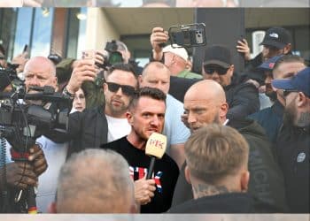 Tommy Robinson in a crowd speaking into a microphone, surrounded by people and cameras, before the riot kicked off