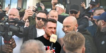 Tommy Robinson in a crowd speaking into a microphone, surrounded by people and cameras, before the riot kicked off