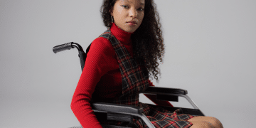 A disabled teenage girl, wearing lipstick to match her autumnal high neck top and tartan dress, looks at the camera with a neutral expression from her wheelchair in a studio